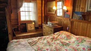 Ein Schlafzimmer in einer Holzhütte mit einem Bett, einem Schminktisch und zwei Fenstern mit Vorhängen.