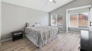 Une chambre à coucher comprenant un lit, une table de chevet, un ventilateur de plafond et deux fenêtres équipées de stores.