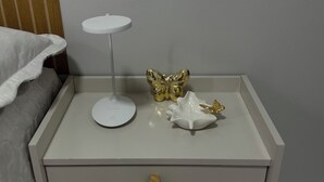 Una mesita de noche blanca con un cajón, una lámpara pequeña blanca y dos adornos dorados.