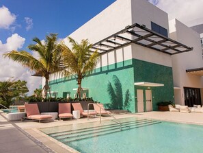 Ein Poolbereich mit Liegestühlen, einer Palme und einem Gebäude mit grün-weißer Fassade.