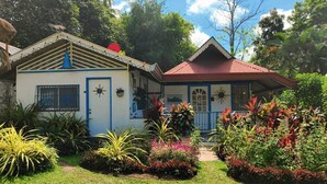 Un edificio de dos pisos con fachada azul y blanca, techo rojo y un jardín con plantas tropicales.