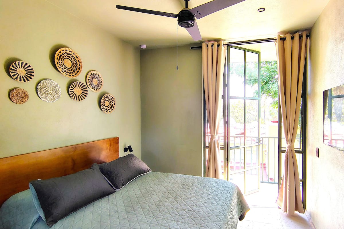 Un dormitorio con cama, ventilador de techo, televisor montado en la pared y ventana grande con cortinas.