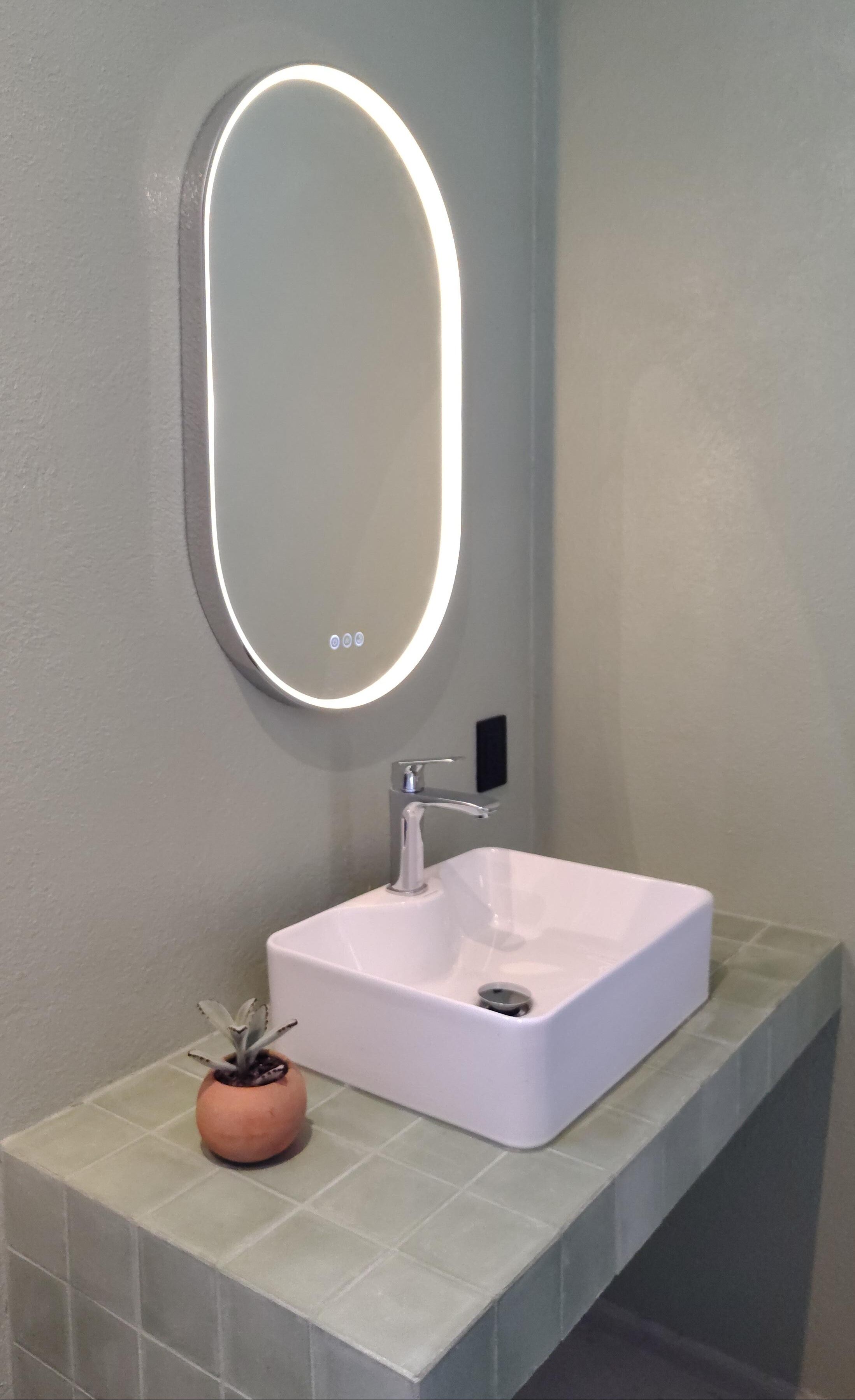 A bathroom with a white sink, a round mirror with lights, a small potted plant, and a tiled countertop.