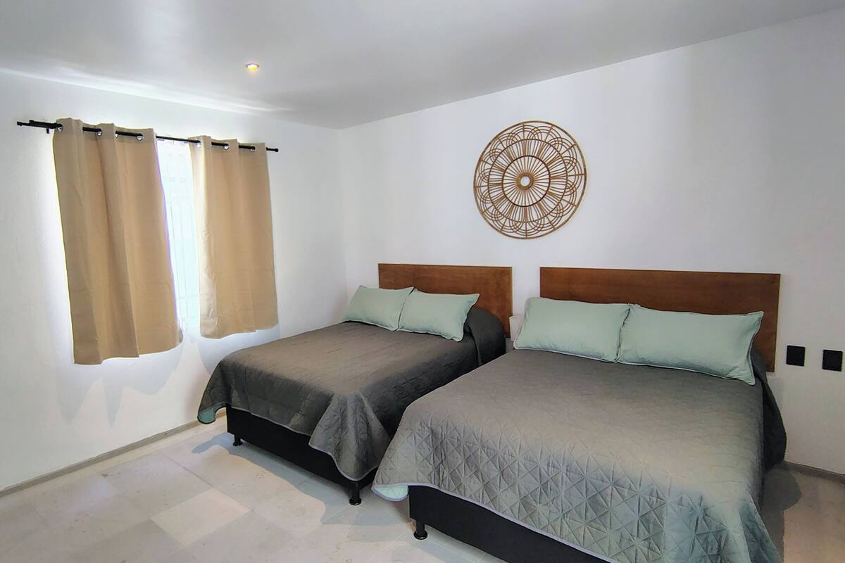 Un dormitorio con dos camas, cortinas color beige, un adorno en la pared y piso de tono claro.