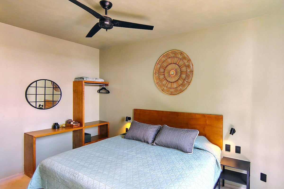 Un dormitorio con cama, ventilador de techo, espejo circular fijado a la pared, mesita de noche de madera y un adorno colgante en la pared.