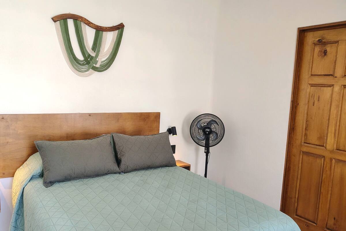 Una cama con cabecera de madera, dos almohadas, una colcha acolchada, una lámpara de pared, un ventilador de pie y una puerta de madera.