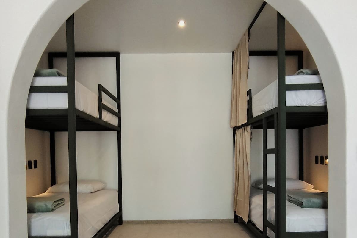 Una habitación con literas bajo un techo abovedado, suelo de baldosas y una lámpara pequeña en el techo.