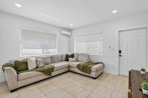 Ein modernes Wohnzimmer mit einem beigefarbenen Ecksofa, großen Fenstern und einer weißen Tür.