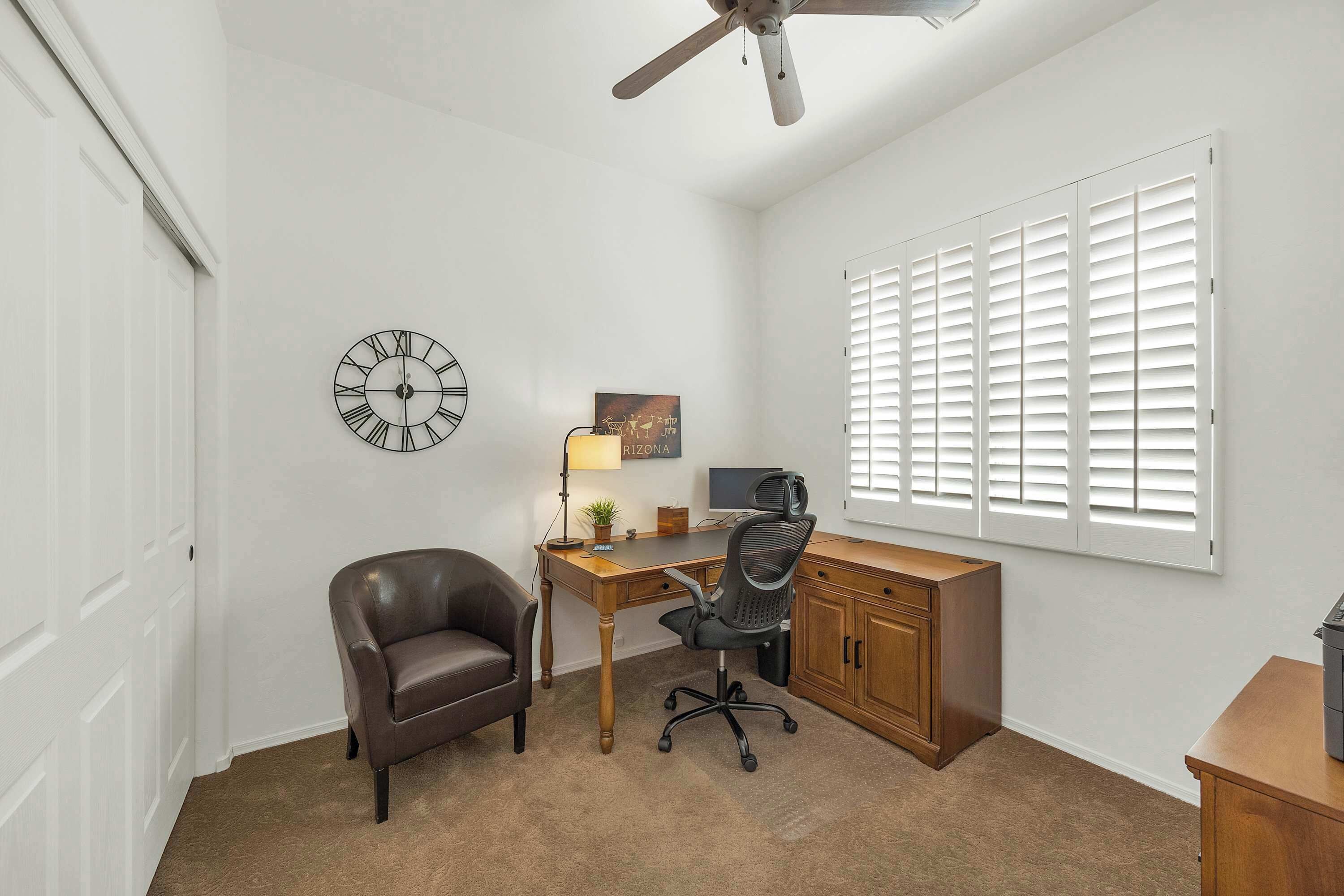 A room with a desk, chair, clock, and window with blinds.