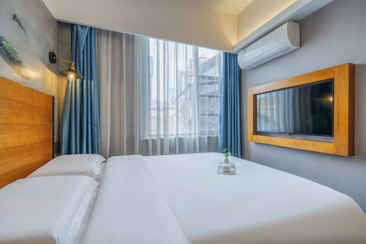 A hotel room with a bed, a wall-mounted TV, an air conditioning unit, and a window with curtains.
