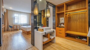 A hotel room with a wooden floor, a bed, a desk with a chair, a vanity with a mirror, and a walk-in closet with shelves. - Gongxili - Chujian Hotel (Kunming)