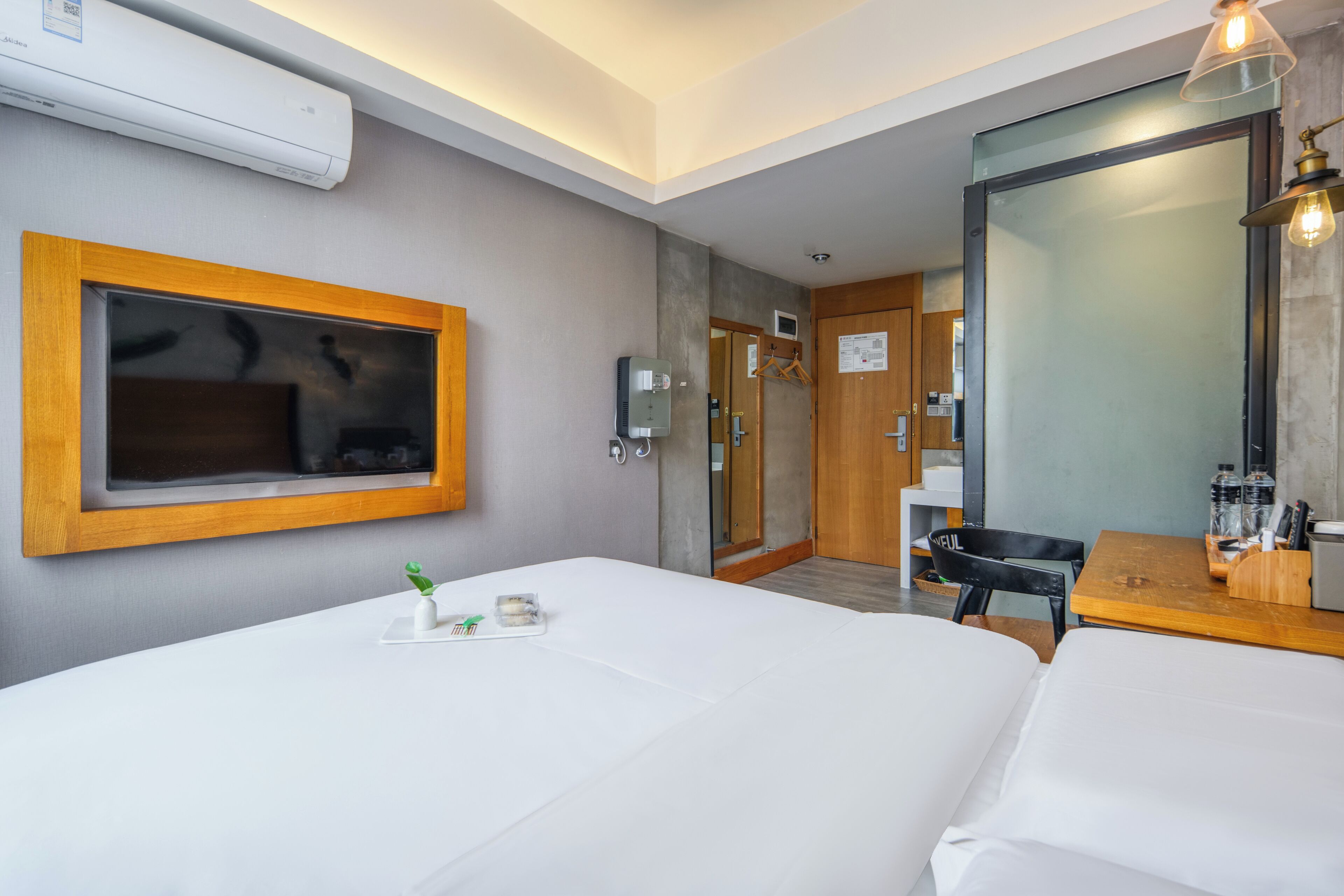 A hotel room with a bed, a desk, a chair, a mirror, and an air conditioning unit.