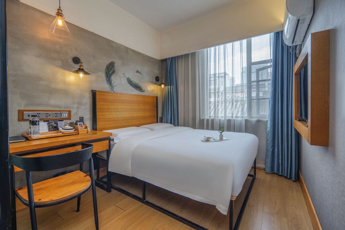 A hotel room with a bed, desk, chair, and a view of the city through the window.