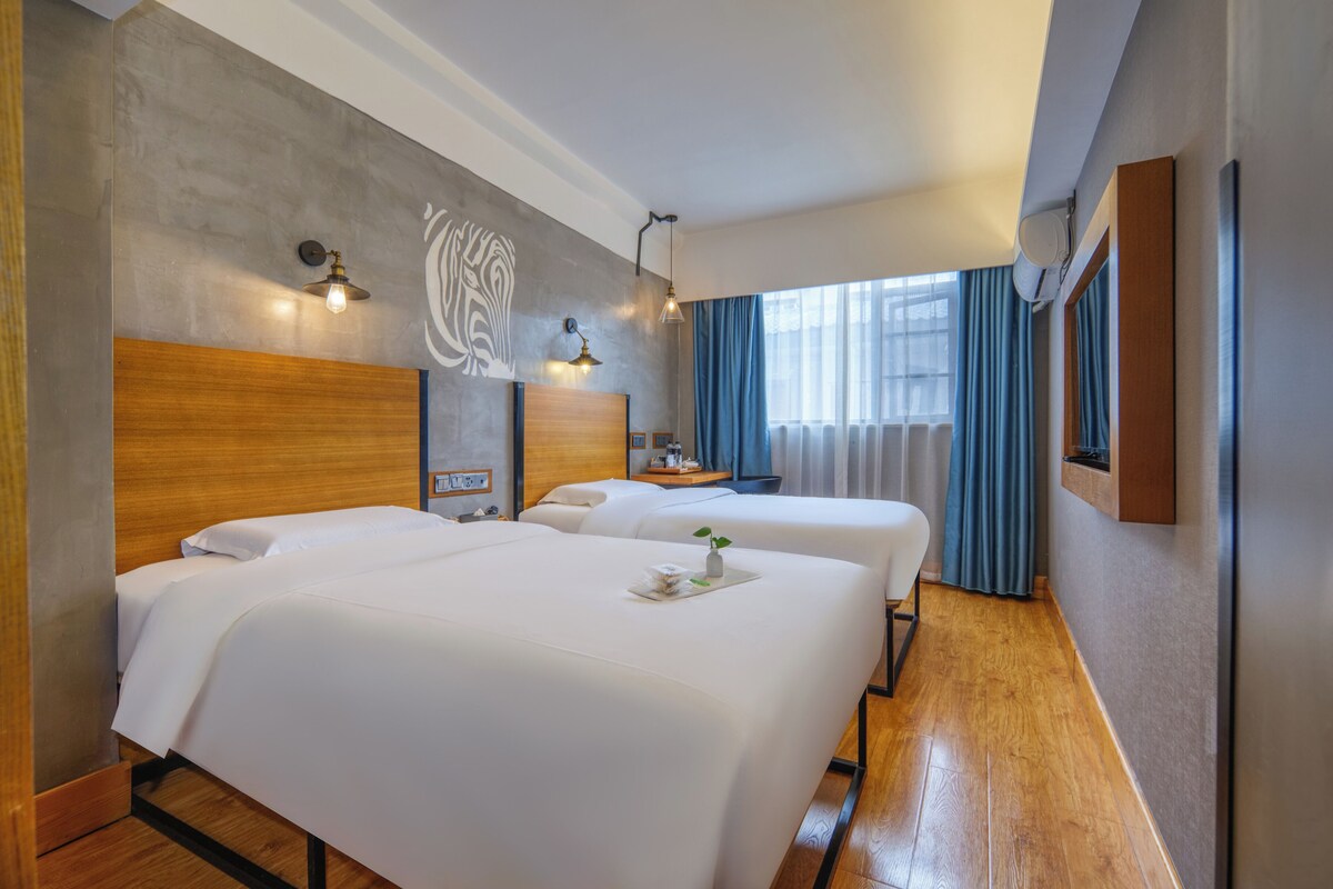 A hotel room with two beds, a wooden headboard, a wall art piece, and a window with curtains.