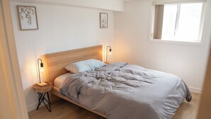 A bedroom with a wooden headboard, a bed with grey bedding, two bedside lamps, and a window with curtains. - Cozy Retro Suite with Garden Walkout, Games & Lounge! (Toronto)