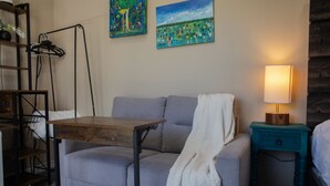 Ein Zimmer mit grauem Sofa, einem Holztisch, einem türkisblauen Nachttisch mit Lampe und zwei Bildern an der Wand.