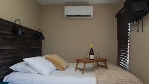 Ein Schlafzimmer mit einem Bett, Kissen, einem Nachttisch mit einer Flasche und Gläsern, einer Klimaanlage und einem Fenster mit Jalousien.