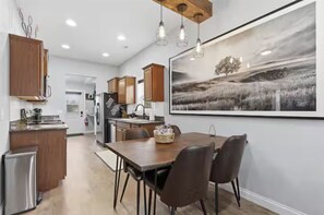 A modern kitchen with wooden cabinets, a dining table with chairs, and a large landscape painting on the wall. - WyNot! Monument House (Rapid City)