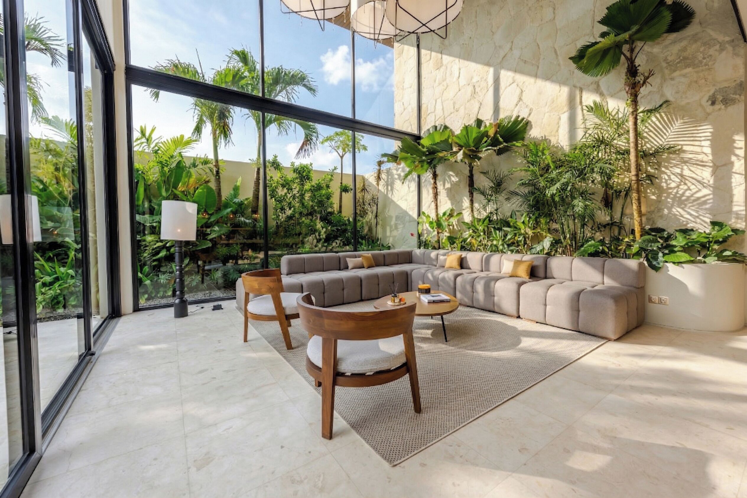 Un salon moderne avec un canapé d’angle gris, des chaises en bois et une table basse. De grandes fenêtres offrent une vue sur un paysage verdoyant.