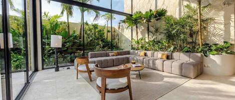 Un salon moderne avec un canapé d’angle gris, des chaises en bois et une table basse. De grandes fenêtres offrent une vue sur un paysage verdoyant.