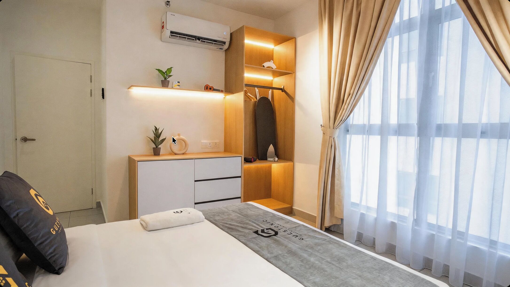 A modern hotel room with a bed, a bedside table, a wardrobe, and a large window with sheer curtains.