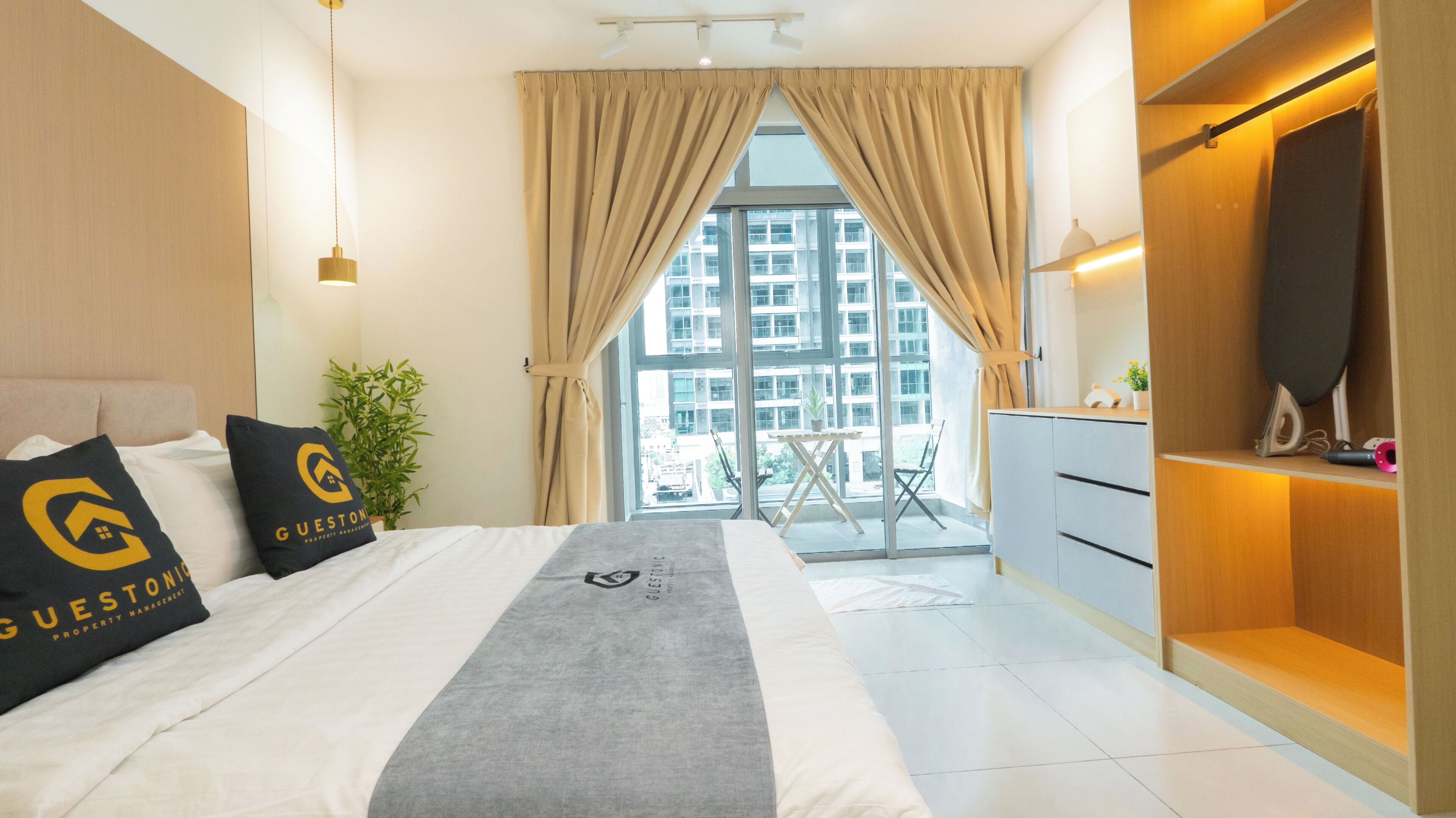 A hotel room with a bed, a desk, a chair, a potted plant, and a view of buildings through the window.