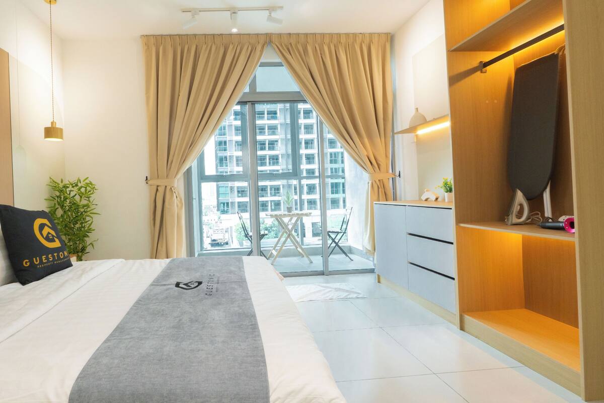 Quarto de hotel com uma cama, uma escrivaninha, uma cadeira, um vaso de planta e vista para edifícios pela janela.