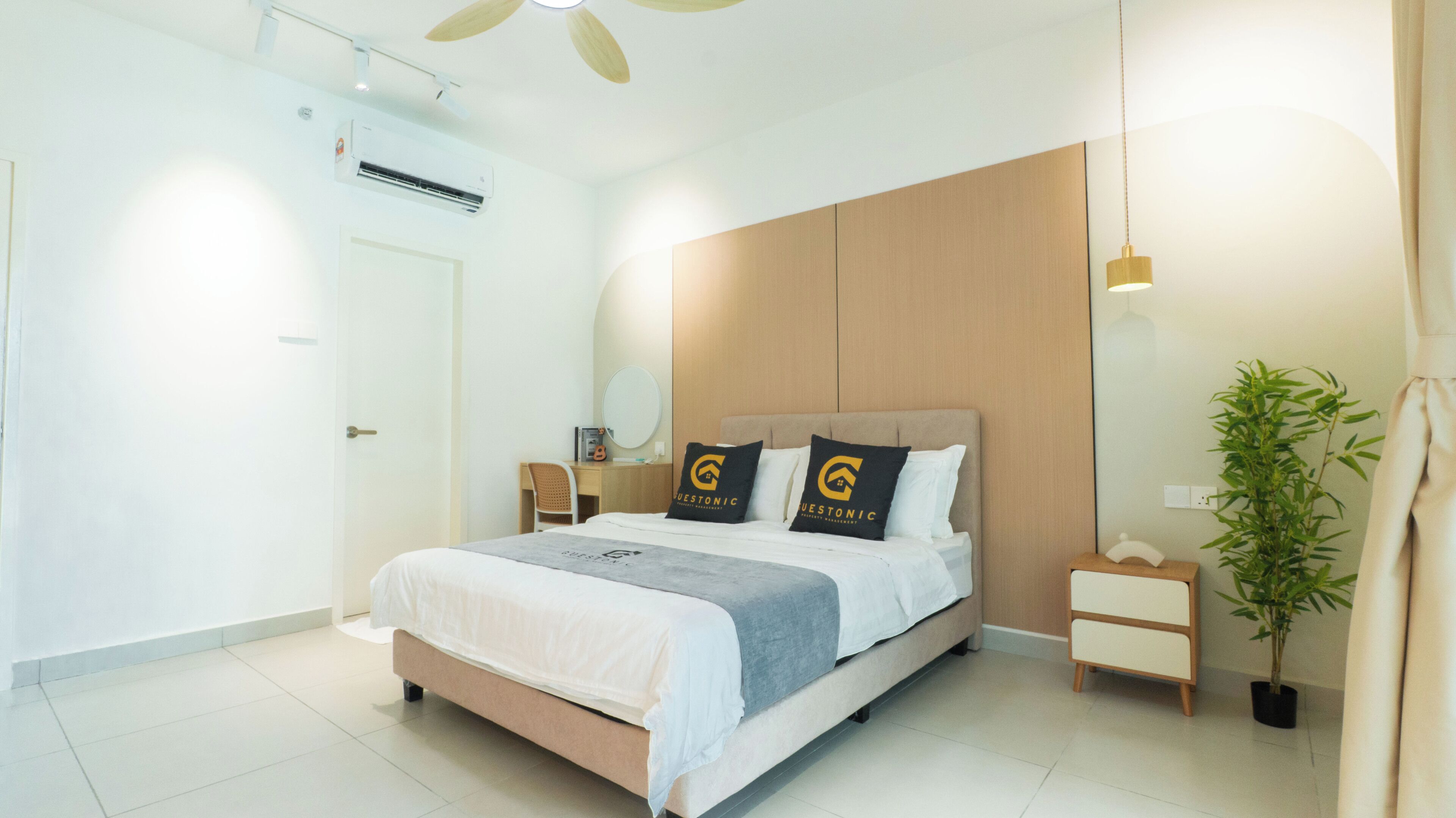 Ein modernes Hotelzimmer mit Bett, Nachttischen, Schreibtisch, Stuhl, Klimaanlage, Deckenventilator, Topfpflanze und Spiegel.