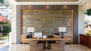 A reception area with a large stone wall, a reception desk, two chairs, a laptop, a calendar, and a coffee machine. - The Gantari Ubud Bali (Ubud)
