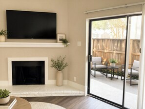 A living room with a fireplace, a flat-screen TV mounted above it, and a sliding glass door leading to an outdoor patio area with a wooden fence and outdoor furniture.