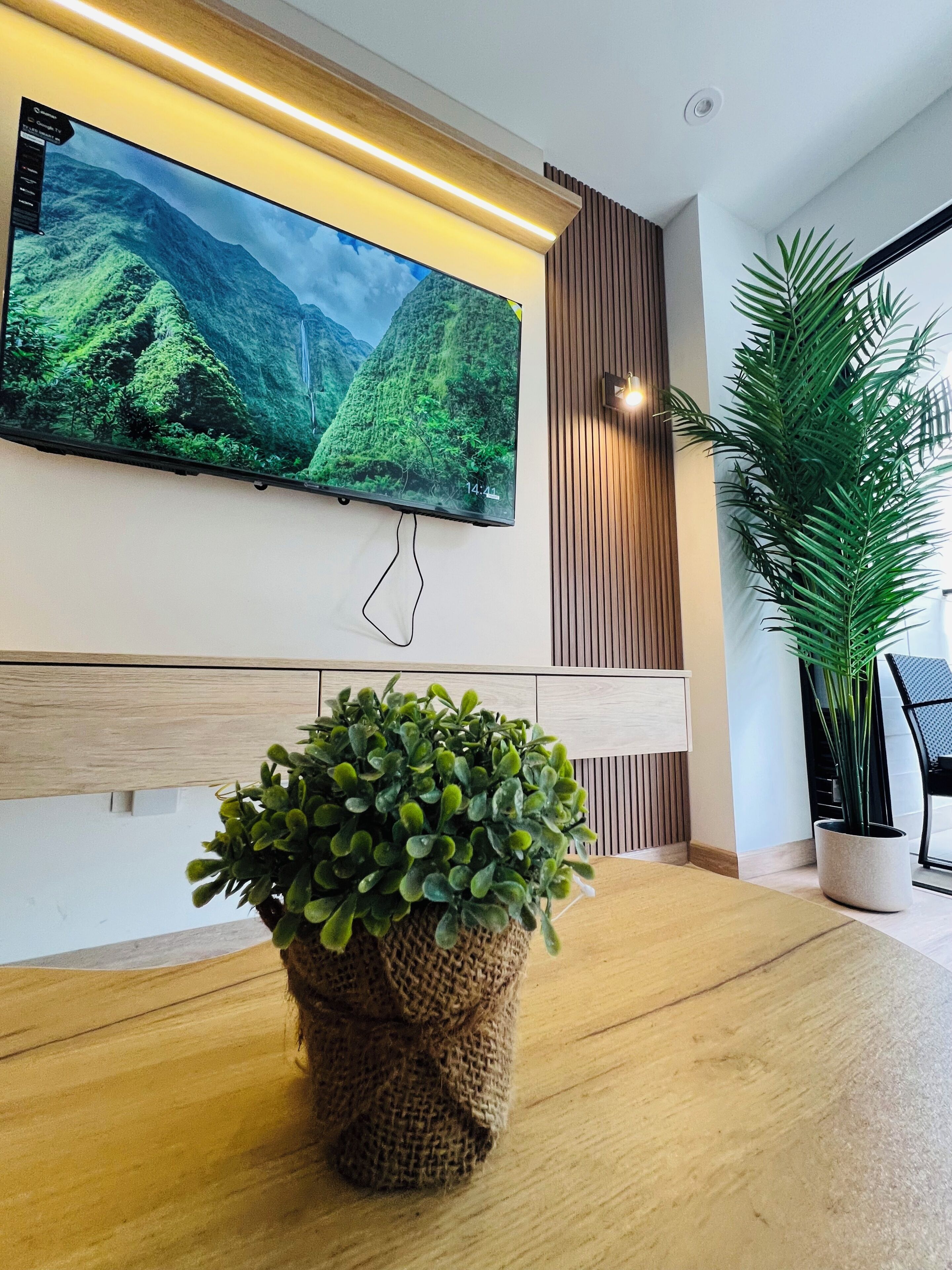 Un interior moderno con un televisor de pantalla plana grande montado en la pared, una mesa de madera y una planta en maceta.