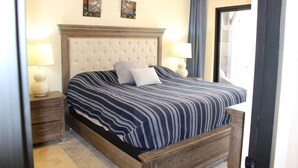 A bedroom with a tufted headboard, a striped bedspread, two bedside lamps, and a nightstand. - 3-Bedroom Villa - Quivira Los Cabos (Cabo San Lucas)