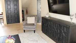 A living room with a grey armchair, a dark wooden console, a flat-screen TV, and a patterned rug. - 3-Bedroom Villa - Quivira Los Cabos (Cabo San Lucas)