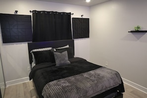 Un dormitorio con cama, cortinas negras, cabecero y estante de pared con una planta.