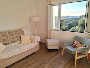 A living room with a sofa, a chair, a small ottoman, a floor lamp, and a window with a view of greenery and buildings. - Can Andreu II apartment next to the beach (Cala Galdana)