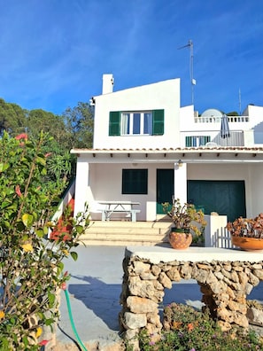 A two-story white building with a balcony, a stone wall, and potted plants. - Can Andreu II apartment next to the beach (Cala Galdana)