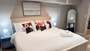 Un letto rifatto con lenzuola bianche, un comodino blu con una lampada, uno specchio grande e un quadro appeso al muro.