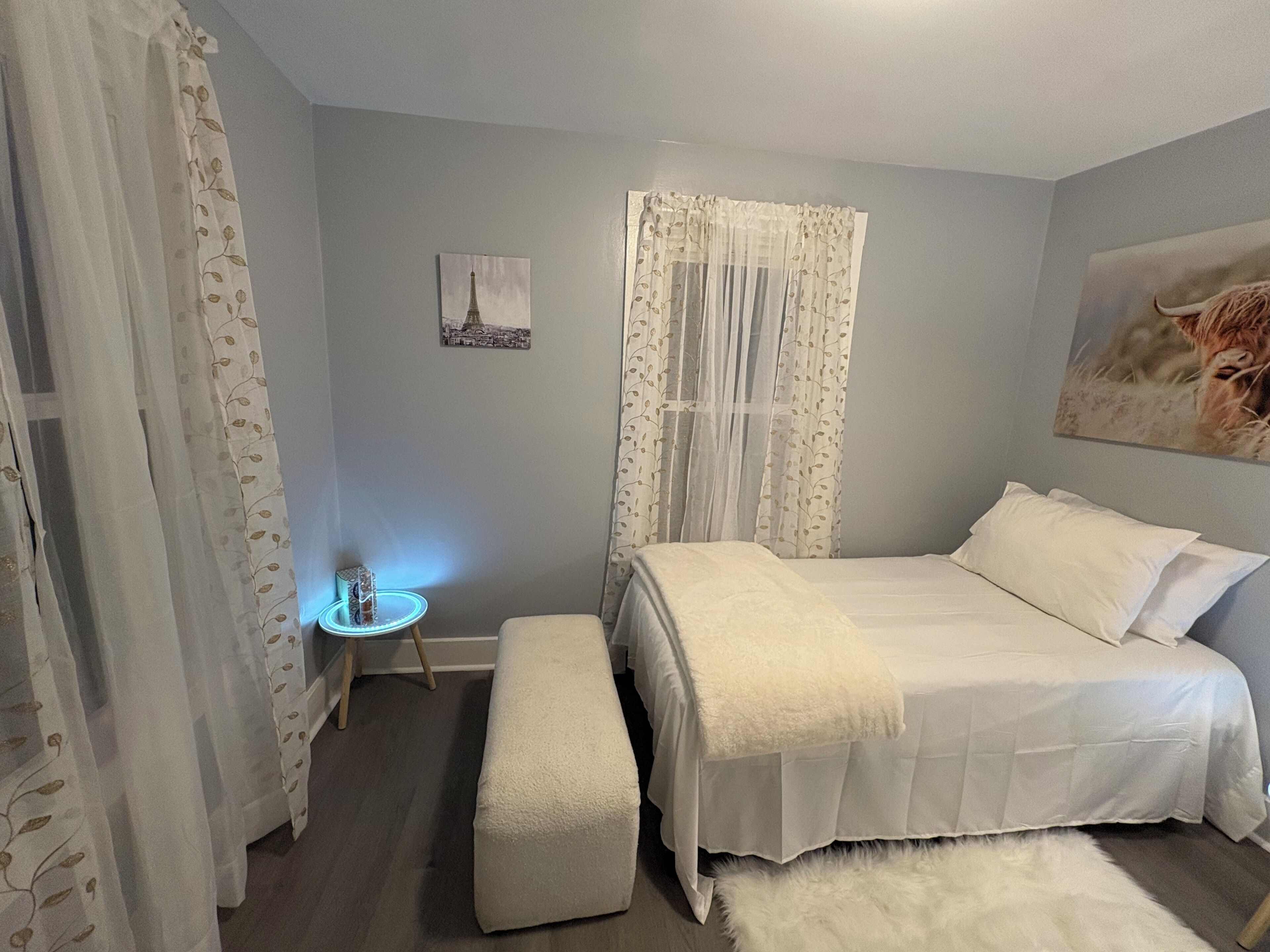 Un dormitorio con cama, una banca, una mesita de noche con lámpara y un cuadro en la pared.