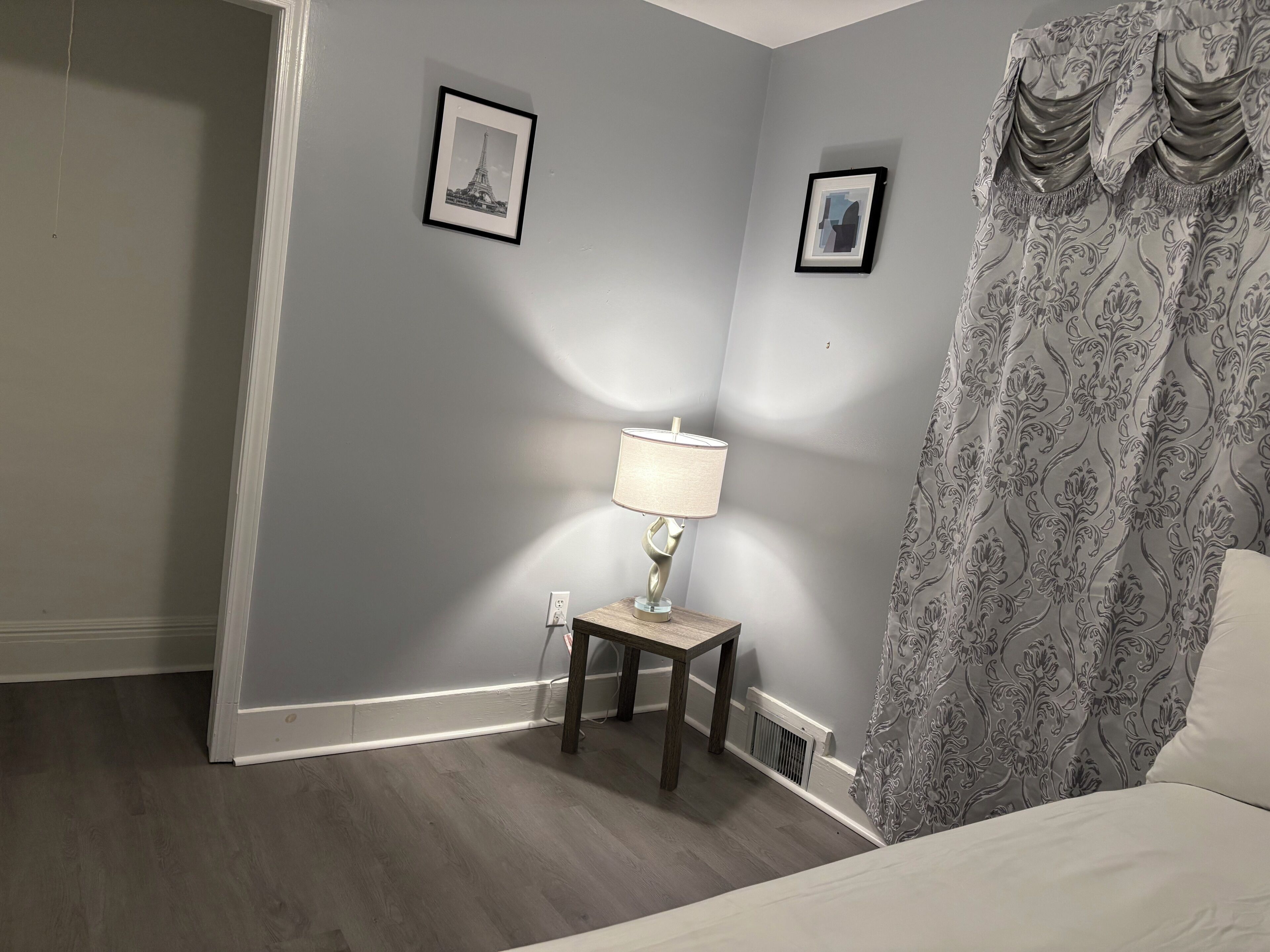 Una habitación con cama, mesita de noche con lámpara, cuadro enmarcado en la pared y cortina estampada.