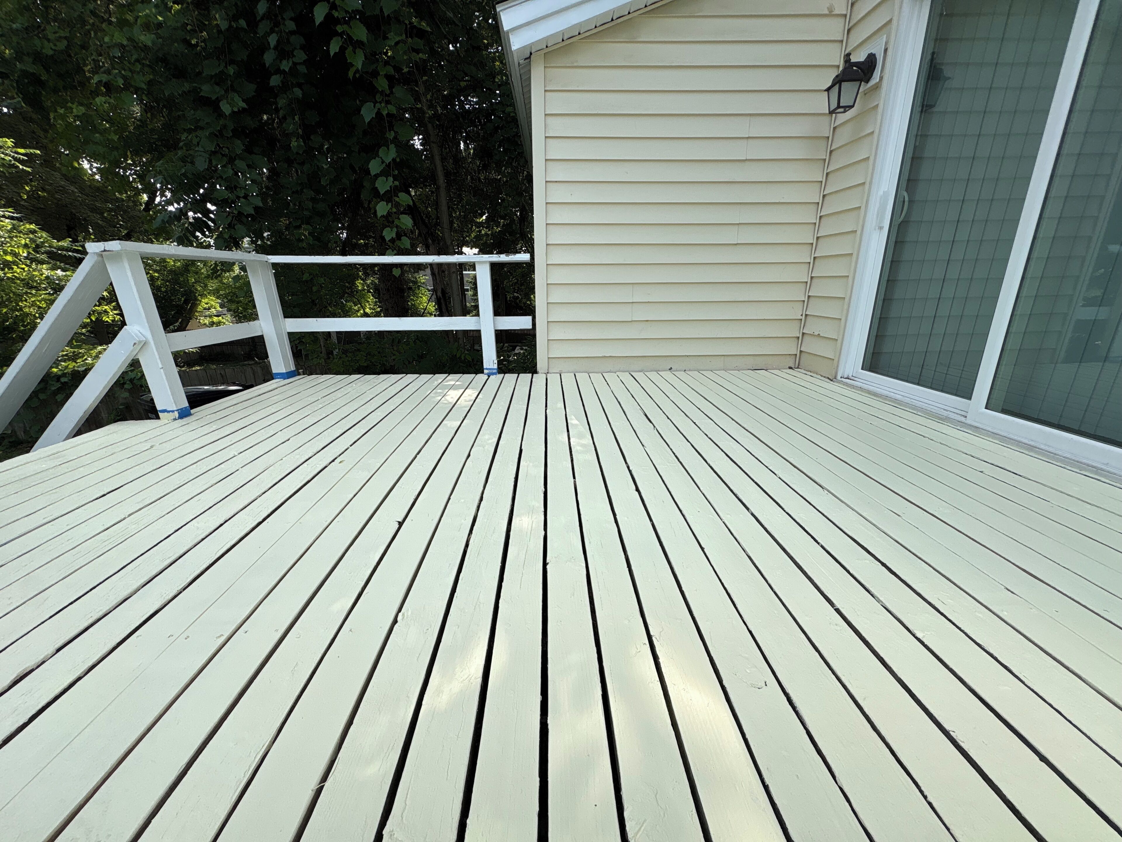 Een houten terras met witte reling, een beige huis en een raam met een rasterpatroon.