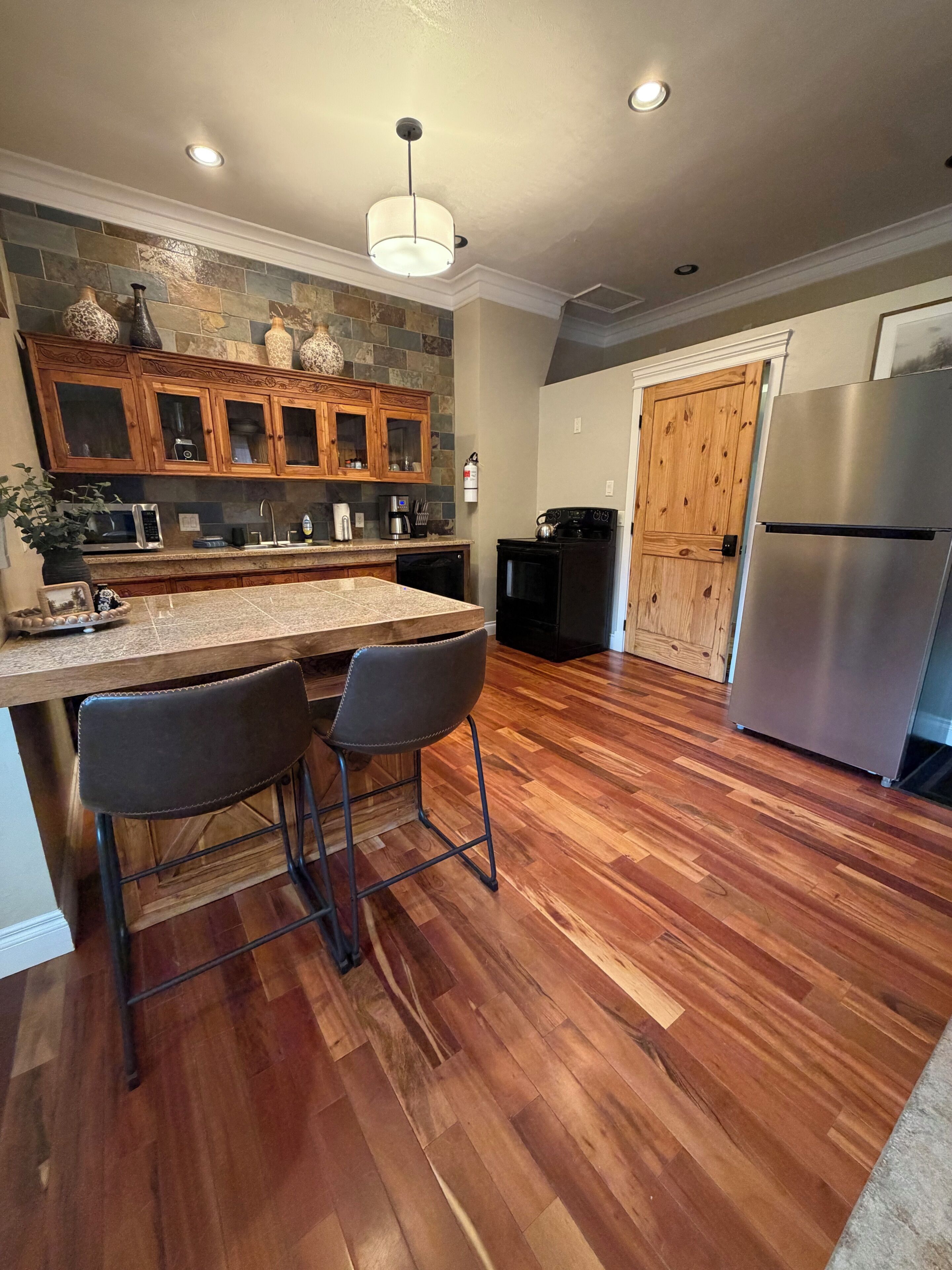 A kitchen with a wooden island, two bar stools, a stainless steel refrigerator, and a stone backsplash.