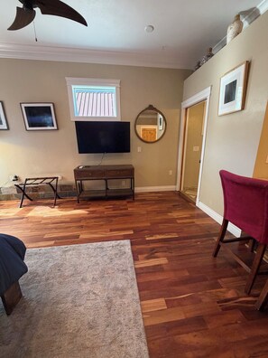 Una sala de estar con piso de madera, un televisor de pantalla plana, un banco de madera, una silla roja y un ventilador de techo.