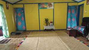 A room with a yellow wall, a bed with a patterned bedspread, a small table with a plant, and a window with curtains.