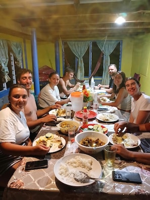 Eine Gruppe von Menschen speist an einem Tisch mit verschiedenen Gerichten, darunter Reis und Salat, in einem Raum mit blauen und gelben Wänden.