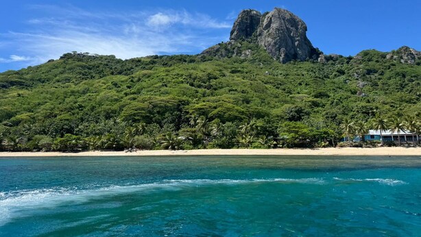 A beach with a prominent rocky mountain in the background and clear blue water.