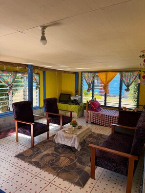 Ein Zimmer mit Deckenventilator, zwei Stühlen, einem kleinen Tisch mit Tischdecke und einem Fenster mit Vorhängen.