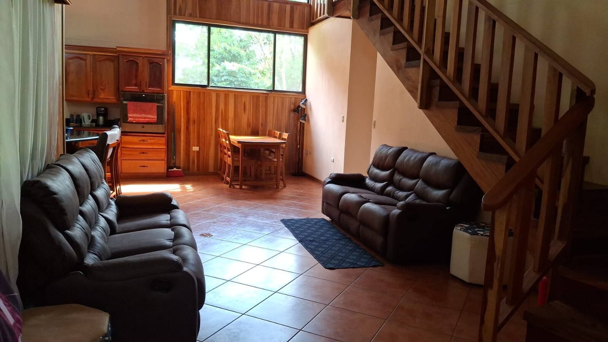 A spacious living area with a dining table, two leather sofas, a staircase, and wooden cabinetry.