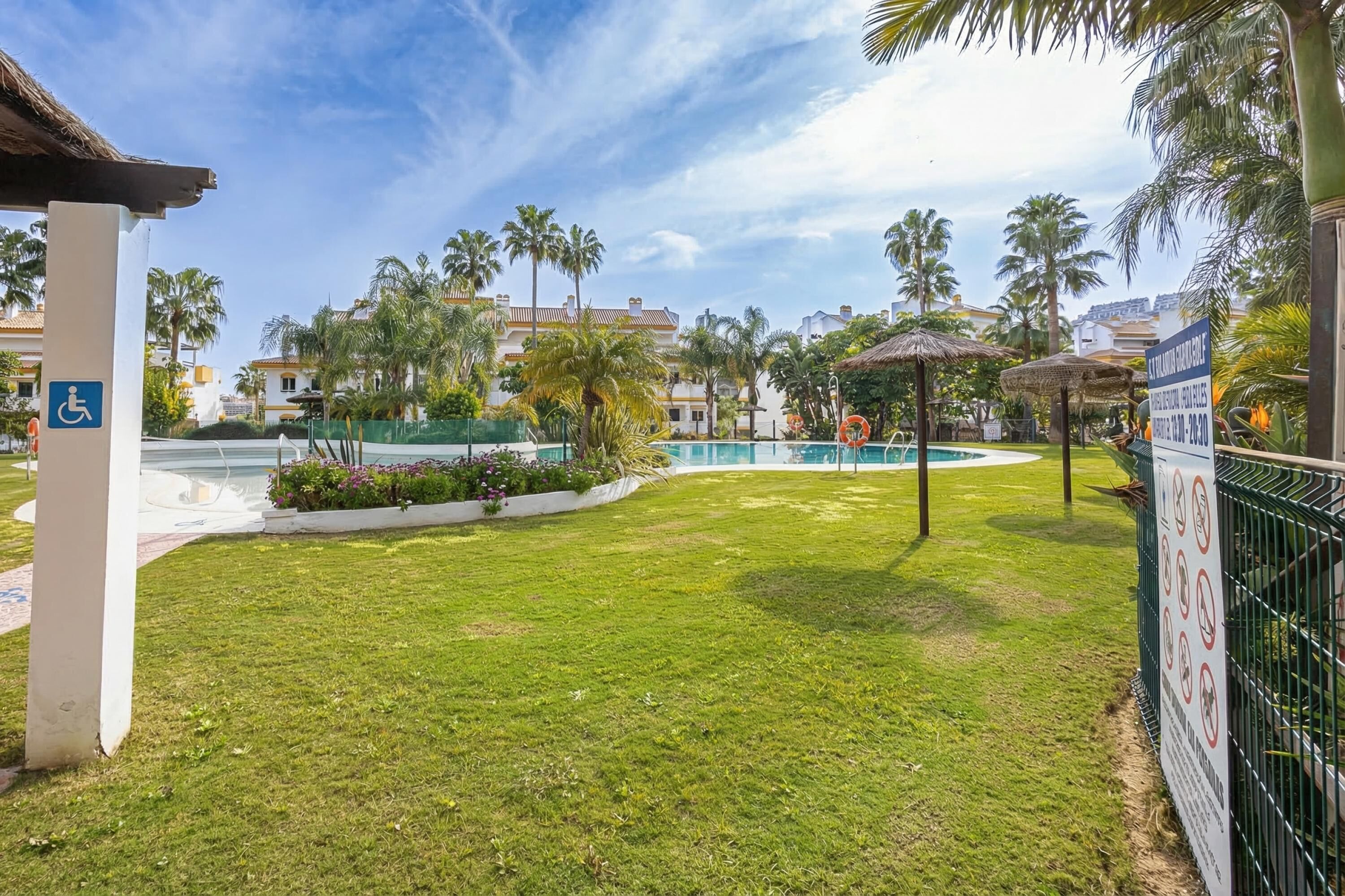 A pool area with a thatched gazebo, palm trees, and a wheelchair-accessible sign.