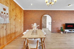 A dining area with a wooden table and chairs, a brick wall, and a TV displaying 'NETFLIX'. - Charming Family Retreat with Arcade + Walk to Shops - Glenunga (Glenunga)