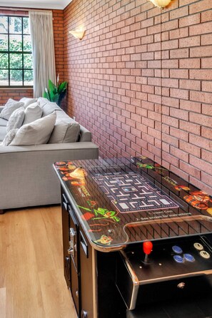 A room with a brick wall, a sofa with cushions, and a table with a game. - Charming Family Retreat with Arcade + Walk to Shops - Glenunga (Glenunga)